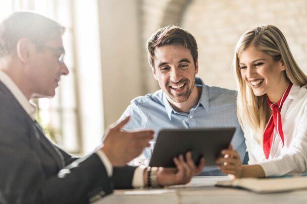 Smiling man and woman looking at computer tablet while a man in a suit explains something to them