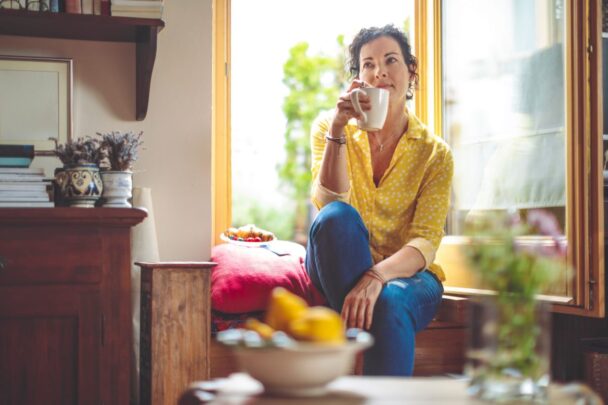 Pensive looking woman holding coffee cup to her lips while sitting in an opened window sill