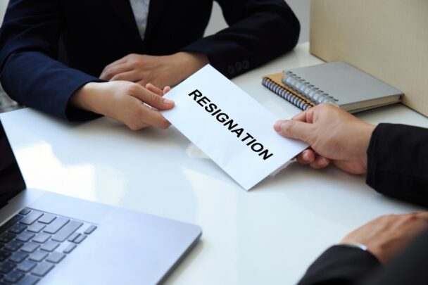 zoomed in perspective of two people sitting at a table exchanging a piece of paper that reads "resignation"