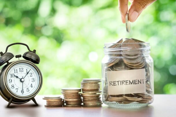 green background with alarm clock, various stacks of coins, and a jar filled with coins labeled "retirement" on a table