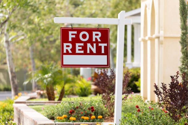 Red sign that reads "for rent" hanging from white post in landscaped front yard
